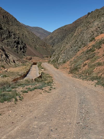 The image depicts a steep mountain landscape. A dirt path runs along the right side, and a concrete aqueduct is visible below. The mountainside is rugged and covered in sparse vegetation. The sky is clear and blue. The location is Krasnyy Most, Kyrgyzstan.
