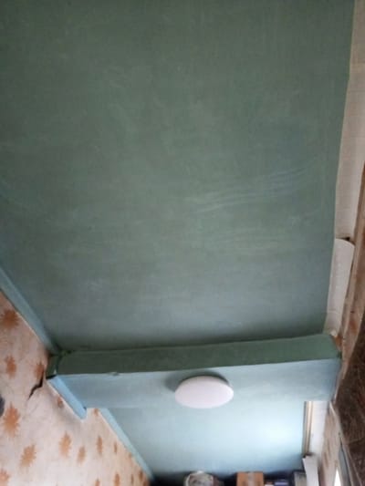 The image shows an interior view of a room, likely in a residential building. The ceiling is painted a muted green color, with a lower section featuring a recessed light fixture. The walls are covered with wallpaper that has an orange floral pattern. One corner of the wall shows signs of damage, with cracks visible in the wallpaper. The bottom of the image shows some obscured items, which might be storage boxes and other household objects. The location in the city of Parabel, Russia, cannot be confirmed or denied from the image alone.