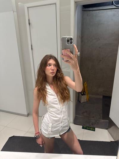 A young woman with long brown hair takes a selfie in a bathroom. She is wearing a white sleeveless top and denim shorts. She holds a smartphone with a pink case. The bathroom has white walls and tiles and a concrete ceiling. A yellow "Caution Wet Floor" sign is visible in the reflection of the mirror. Based on the surroundings, the location is identified as a bathroom in the Hongkou District, China.