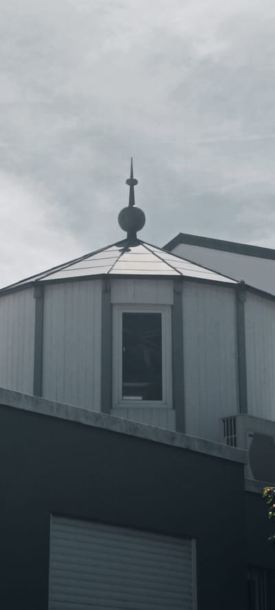 The image shows the upper portion of a building, specifically a domed structure with a window and a spire on top. The building is painted white with gray trim. Below the domed structure is a gray building with a closed roll-up door. In the background, a cloudy sky is visible. The location is identified as Winterbach, Germany. There are no people or animals visible in the image. The scene appears to be a static architectural view.