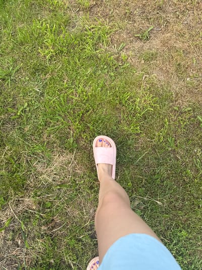 The image shows a close-up, high-angle view of a person's left leg and foot standing on a grassy area. The person is wearing a light blue garment, possibly shorts or a skirt, and a pink slide sandal on their left foot. The sandal reveals toes painted with blue nail polish. The ground is covered in a mix of green and dry grass, with some patches of bare earth. The overall scene suggests an outdoor setting. The location is described as Луптюг, Russia.