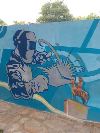 The image shows a mural on a blue wall. The mural depicts a welder wearing a welding mask and holding a welding torch, with sparks flying from the torch. To the right of the welder, a figure of a person riding a horse is painted on top of what seems to be a pedestal. The environment around the wall appears to be an outdoor setting with some vegetation visible in the background. The city of Villa Hayes, Paraguay, is mentioned as the location of the image.