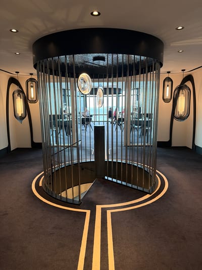 The image depicts an interior scene, likely a modern staircase or atrium, characterized by its sleek and contemporary design. The dominant elements are a cylindrical structure wrapped in horizontal metallic bars, creating a sense of depth and reflection. Within this structure, the blurry presence of people can be seen, suggesting a public or commercial space, possibly a hotel, office building, or restaurant. They appear to be seated at tables, engaged in quiet activities.
The lighting is a prominent feature, with several modern pendant lights featuring elongated, tubular bulbs and decorative, cage-like shades hanging from the ceiling. Recessed spotlights are also visible on the white walls. The floor is carpeted in a dark gray material, with a distinctive curving design in a lighter color, possibly cream or beige, accentuating the architectural lines of the space.
The overall aesthetic is sophisticated and minimalist, with a focus on geometric shapes and reflective surfaces. The lighting creates an ambiance that is both elegant and functional. The time of day is suggested to be daytime, given the natural light filtering through unseen windows, though the specific weather conditions are not discernible. The scene conveys a sense of calm and order. No visible text is present in the image. The location context of La Clarté, France, suggests a European setting for this modern interior.
