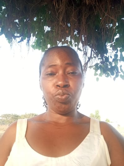A woman with dark skin puckers her lips toward the camera. She wears a white tank top and dangling earrings. Above her head, a tree with hanging roots and green leaves creates a canopy. A field with sparse vegetation stretches out in the background under a bright sky. The light is harsh, creating shadows on her neck and face.