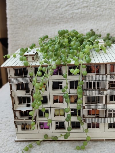 A still life image features a miniature wooden model of a Soviet-era apartment building, possibly located in Novomoskovsk, Russia. The model is intricately detailed, showcasing multiple floors with small, square windows, some of which appear to have colorful interior lighting. A string of pearls succulent plant drapes over the roof and cascades down the side of the building, its round, green leaves resembling pearls. The model and plant are placed on a textured, light-colored surface, possibly a rug or blanket. The background is a soft, out-of-focus texture, also light in color. There are no people or animals present in the image.