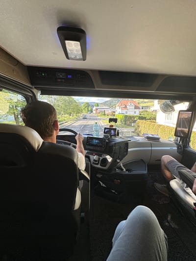 The image shows an interior view from a vehicle moving along a road in what appears to be Sankt Marein bei Neumarkt, Austria. The driver, a man with short brown hair, is behind the steering wheel. A passenger is seated to the right of the driver. In the background, outside the vehicle's windshield, is a street view with buildings, trees, and other vehicles. A navigation system and other electronic devices are visible on the dashboard. A light and control panel are attached to the ceiling or a shelf above the dashboard.