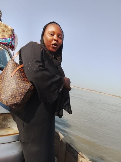 The image depicts a daylight scene on a body of water in Ibi, Nigeria, likely a river crossing. A female individual, an adult with light brown complexion, is positioned in the left-center foreground, standing on what appears to be a wooden boat or ferry. She wears a black abaya with a dark head covering; the sleeves of her garment show scattered light-colored embellishments. A brown shoulder bag with a repeating geometric pattern and tan straps is carried on her left side. Her face is directed towards the viewer, with her mouth slightly open. To her immediate left, the front portion of a light-colored vehicle, possibly a minivan or bus, is visible, including a headlight and part of the bumper/hood. Luggage covered in patterned fabric is secured on the vehicle's roof. The right side of the frame features a wide expanse of light grey-blue water with minor ripples. A faint, distant shoreline with sparse vegetation is visible across the water, meeting a clear, light blue sky above.