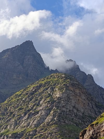 This is a close-up, low-angle shot of jagged, rocky mountains in Damphug, India. The peaks are sharp and imposing, with dark, textured surfaces. Some of the lower slopes are covered in patches of vibrant green vegetation, contrasting with the gray rock. Wisps of white clouds drift across a bright blue sky, partially obscuring some of the higher mountain peaks, creating a dramatic and somewhat mysterious atmosphere. The image focuses on the rugged, natural beauty of the mountainous terrain.