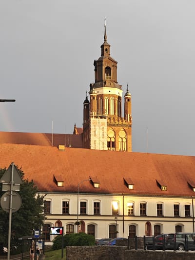 The image captures a view of a building with a prominent clock tower in Stargard, Poland. The scene depicts an urban setting with architectural elements. In the foreground, there's a road with several parked cars, bordered by a low stone wall. To the left, a bus stop with signage and two young women can be seen. Behind them, a building with an orange tiled roof stretches across the frame, featuring multiple windows and dormer windows. The architecture of this building suggests it's of historical significance.
Dominating the upper portion of the image is a tall, ornate clock tower made of brick. The tower has multiple levels, decorative windows, and a spire topped with a cross. A large banner with text is draped across a section of the tower, indicating ongoing construction or renovation. The text on the banner reads "quick-mix".
The time of day appears to be late afternoon or early evening, as evidenced by the warm sunlight reflecting off the windows of the building and the golden hue on the tower. The sky is a pale grey, suggesting a slightly overcast or hazy atmosphere. The overall mood is tranquil, with a focus on the imposing architecture and the interplay of light and shadow. The location cues like the bus stop and the style of the buildings confirm the urban context of Stargard, Poland.