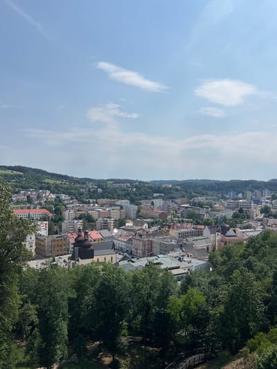 The image presents an elevated view of the city of Náchod, Czechia. The foreground is dominated by lush green trees, obscuring the lower portion of the cityscape. Beyond the trees, a dense urban area unfolds, characterized by a mix of multi-story residential buildings and older architectural structures. Prominent among these buildings is a church with two dark, domed bell towers. The city extends towards a backdrop of rolling, tree-covered hills under a bright blue sky with scattered clouds. The perspective suggests the photograph was taken from a higher vantage point, possibly a hill or a tall building overlooking the city.