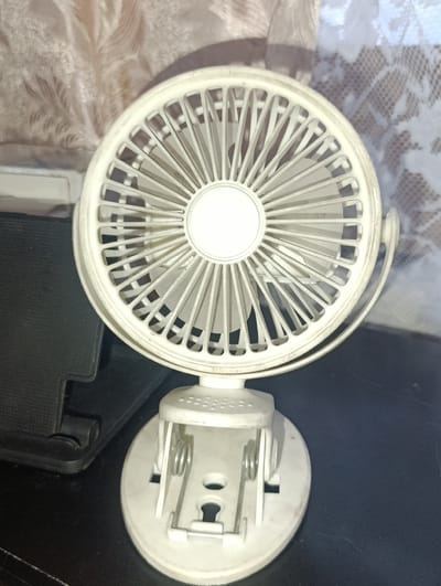 A white, battery-operated fan with a clip-on base is the central subject of the image. The fan head is circular with a protective grille and blades, and it is attached to a white stand with springs and a clip mechanism. The fan is positioned on a dark, textured surface, possibly a table or shelf. In the background, a sheer curtain with a patterned design is visible, with soft light filtering through it. The overall environment appears to be indoors, likely a room. The image does not provide any information or context related to the city of Yenakiieve, Ukraine.