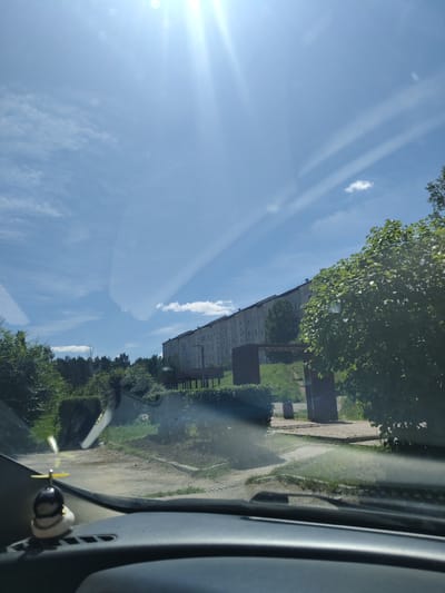 The image captures a view from inside a car, looking out through the windshield. The scene depicts a bright, sunny day with a clear blue sky, punctuated by faint streaks of clouds and lens flare from the sun. In the mid-ground, a long, multi-story apartment building with numerous windows stretches horizontally across the frame. In front of the building, a line of trees and some greenery are visible, with a wooden gazebo-like structure positioned on a slightly elevated platform. To the right, a large, leafy green tree partially obstructs the view of the building. The foreground shows the dashboard of the car, with a small decorative item, resembling a black and white figurine with a yellow propeller, positioned on the left side. The ground in front of the car appears to be a dirt or gravel surface with some patches of grass. The overall environment suggests a residential area on the outskirts of a city, possibly Shimanovsk, Russia, given the architectural style and surrounding vegetation.