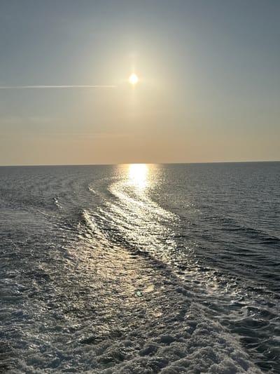 The image depicts an expansive open sea under a clear sky, located in the vicinity of Palaiopoli, Greece. The sun, a bright, starburst-like point of light, is positioned in the upper-left quadrant, low enough to create a pronounced, shimmering reflection path extending across the water from the horizon towards the foreground. A faint, linear contrail is visible horizontally in the upper-left sky. The sky transitions from a pale blue at the top to a hazy golden hue closer to the horizon. The lower portion of the frame prominently features a wide, frothy white wake, indicating the recent passage of a watercraft moving away from the viewer. The water outside the direct sun reflection and wake appears dark blue-green with subtle ripples. No individuals, landforms, or artificial structures are visible.