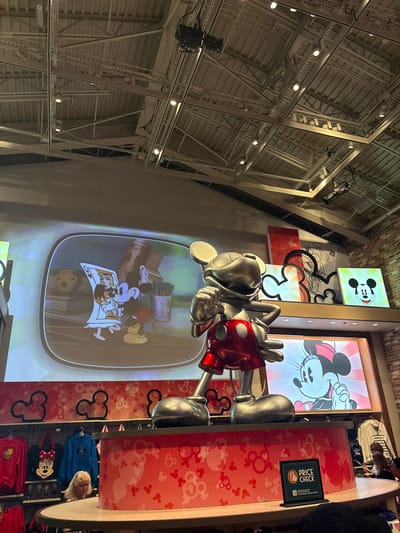 The image depicts a large, shiny metallic statue of Mickey Mouse standing on a raised red display platform. The statue is in a dynamic pose, with one arm bent and the other extended. Behind the statue, a large screen displays animated Disney characters. To the left, a portion of the screen shows Mickey Mouse holding a playing card with the Queen of Hearts. To the right, a framed picture shows Minnie Mouse smiling. Above these displays, a wall features several artworks of Mickey Mouse's ears and face in various artistic styles.
The environment appears to be an indoor retail space, possibly a Disney store, given the prominent display of Disney characters and merchandise. The ceiling shows exposed industrial beams and lighting fixtures. Below the statue, shelves are visible, stocked with Disney-themed apparel, including t-shirts and bags. A woman with blonde hair is partially visible behind the display platform, looking towards the merchandise. Another woman is seen in the background, interacting with merchandise. A "PRICE CHECK" sign is placed on the display platform. The overall setting suggests a vibrant, commercial atmosphere dedicated to Disney merchandise in Lake Buena Vista, Florida.