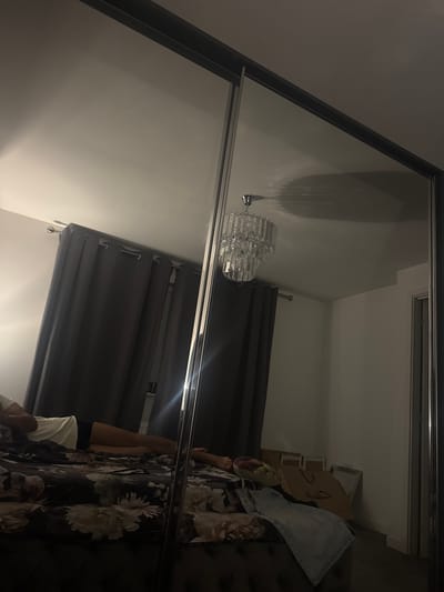 The image shows a bedroom with a mirrored wardrobe reflecting part of the room. A person is lying on a bed with floral bedding. Dark grey curtains are drawn. A chandelier is reflected in the wardrobe mirror. In the background, several cardboard boxes and a framed picture are visible. The location is a bedroom in the city of Peterborough, United Kingdom.