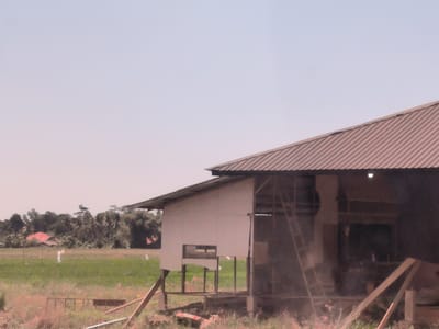 The image shows a partially constructed or dilapidated building in Putoh Sa, Indonesia. The structure has a corrugated metal roof and what appears to be a wooden frame with incomplete walls. A ladder leans against the inside of the building. In the background is a field of vegetation with scattered trees and a glimpse of a red-roofed building in the distance. The foreground shows overgrown grass and construction debris. The overall scene suggests a rural or semi-rural environment.