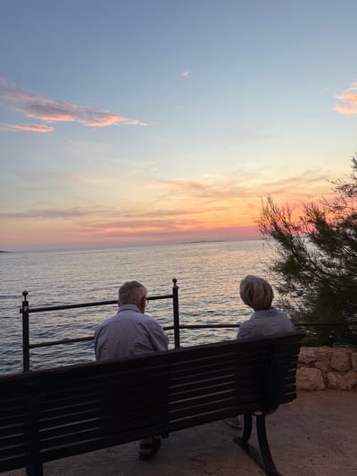 Two elderly individuals are seated on a dark wooden bench facing a body of water, presumably the Adriatic Sea. The man on the left, with gray hair and glasses, wears a light blue collared shirt and dark sandals. The woman on the right has blonde hair and wears a gray top. They are positioned with their backs to the camera, gazing at a sunset.

The sky displays a gradient of colors, transitioning from pale blue at the top to hues of pink, orange, and yellow near the horizon, indicative of a sunset. Wispy clouds are visible in the sky, catching the warm light. The sea is calm, with gentle ripples reflecting the colors of the sky. A dark metal railing runs along the edge of the waterfront in front of the bench. To the right of the bench, a rocky embankment with a shrub is visible.

The bench itself is dark wood with a slatted design and is situated on a paved surface. The scene is set outdoors, likely in a coastal area of Primošten, Croatia, given the characteristic landscape and sea. The overall mood is peaceful and contemplative, with the couple seemingly enjoying the sunset together.