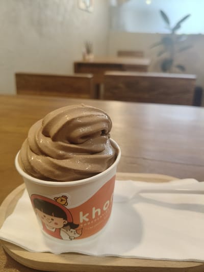 A close-up, eye-level shot focuses on a cup of chocolate soft-serve ice cream. The ice cream is swirled high, with a glossy sheen indicating a cool, creamy texture. The cup features a cartoon illustration of a girl in a red hood, holding a white cat, and a yellow chick above her head. Text on the cup, partially visible, reads "kho" and "CHOCOLATIER." The cup rests on a white napkin, which is on a light-colored wooden tray. A white plastic spoon is placed on the napkin to the right of the cup.
The background is blurred but suggests an indoor setting with wooden tables and chairs. A plant is visible in the upper right corner, and a wall with some subtle textured finish and possibly a framed picture is on the left. The lighting appears to be soft, possibly from natural light filtering in or ambient indoor lighting.
This scene is likely captured within a cafe or dessert shop in Chiang Rai City Municipality, Thailand, given the context of soft-serve ice cream establishments and the typical interior decor of such businesses in the region.
