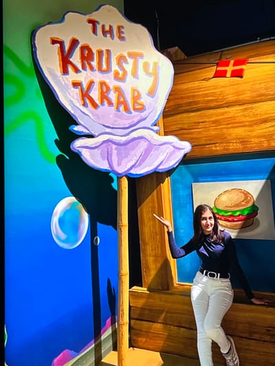 In a playful, cartoon-like setting inspired by SpongeBob SquarePants, a young woman poses near a large, stylized sign that reads "THE KRUSTY KRAB." The sign is shaped like a clam shell and features bold, orange and red lettering with blue outlines. Behind the sign, wooden walls create a backdrop, adorned with a small red and yellow flag.
The woman is dressed in a dark long-sleeved shirt and white pants, with a distinctive white belt. She is leaning against the wall and pointing towards a cartoonish rendition of a Krabby Patty burger, which appears to be embedded in the wall. Her pose and expression suggest she is enjoying the immersive experience.
The scene is set against a bright blue background that evokes an underwater environment, complete with painted bubbles. The overall atmosphere is cheerful and whimsical, designed to resemble a fun attraction or exhibit. The image appears to have been taken indoors, likely in a themed entertainment venue. While the location context is Nijmegen, Netherlands, this is not visually evident in the provided image.