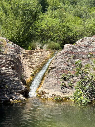 This media file captures a natural scene of a small waterfall cascading over a rocky cliff into a calm body of water. The rocks are a prominent feature, exhibiting a layered texture with striations of reddish-brown and grey, indicating geological stratification. Vegetation, primarily lush green trees and shrubs, thrives on the left side of the frame and also clings to the edges of the rock formations, suggesting a vibrant, possibly humid, environment.
A narrow stream of water is actively flowing down the rock face, creating a foamy white cascade that contrasts with the earthy tones of the stone. The water in the pool below is a greenish hue and shows subtle ripples, reflecting the surrounding light. Some dry grasses and smaller plants are visible growing amongst the rocks and at the water's edge.
The lighting suggests it is daytime, with sunlight illuminating the scene and casting shadows, particularly on the rugged rock surfaces. The overall impression is one of serene natural beauty, likely found in a secluded or less-traveled area. Given the location context of Mazići, Montenegro, this could depict a scene from the region's natural landscape. No people, objects, or text are visible in the image.