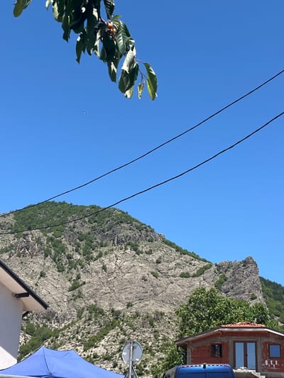 The image is an upward-angled view in Bukovo, Bulgaria, featuring a mountain slope covered with vegetation. A portion of a building with visible windows and a blue van is on the right side of the image. Three power lines span the top of the frame from left to right. The sky is clear and blue. Leaves and branches of a tree or bush are visible in the lower-left corner. Part of a white building with a black roof edge and a blue umbrella is visible in the lower right. A round mirror on a pole is located next to the umbrella.