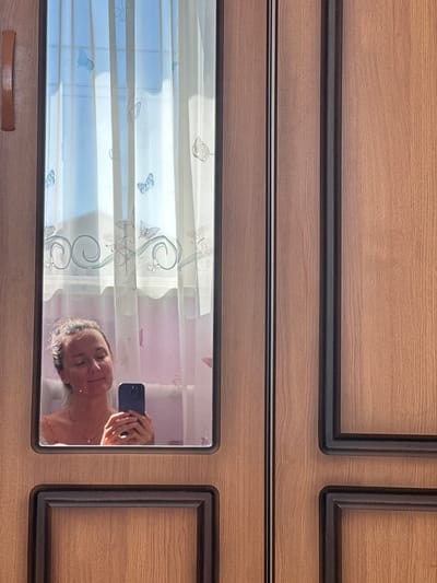 In the city of Rasskazovo, Russia, a woman is reflected in a mirror on a wooden wardrobe. She is holding a smartphone in her hands, seemingly taking a selfie, with her eyes closed and a slight smile on her face. She is wearing a necklace with a pendant and a pink strap visible from her clothing. Behind her, a sheer curtain with a butterfly pattern is illuminated by sunlight. The wardrobe itself has a wood-grain texture and is divided into panels.