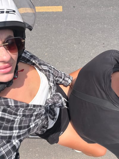In a ground-level, eye-level shot, two individuals are positioned on a paved surface, possibly a road, with a yellow line marking present. The subject on the left, wearing a white helmet with a clear visor, is captured from the chest up, facing slightly to the right. They have on oversized sunglasses with brown lenses and a plaid shirt tied at the waist over a white tank top. Their left arm is visible, partially obscured by the plaid shirt. The individual on the right is seen from the shoulders down, wearing a black t-shirt and black shorts, with their right leg exposed. Their left arm is bent and resting. The overall scene is outdoors, under bright lighting, with the asphalt ground dominating the lower portion of the frame. The image was taken in Dien Tho, Vietnam.