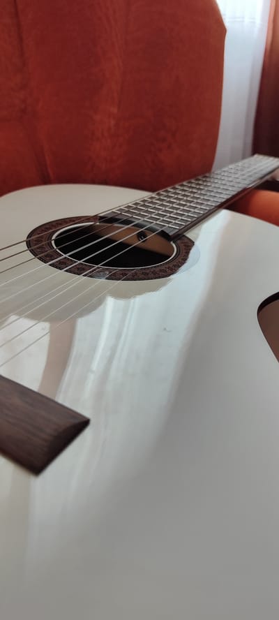 A white acoustic guitar is placed on a surface, with its soundhole and strings visible. The guitar's body reflects light, creating a glossy effect. In the background, a blurred orange surface, possibly a chair or sofa, is visible. To the right, a sheer white curtain hangs, hinting at an indoor setting. The overall scene is still and quiet. This image was taken in the city of Лёзненскі сельскі Савет, Belarus.