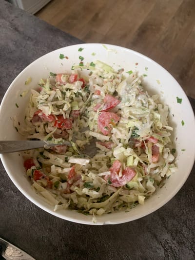A close-up overhead shot shows a white bowl filled with coleslaw. The coleslaw is composed of shredded cabbage, diced tomatoes, and chopped green herbs, all coated in a creamy dressing. A silver fork rests on the edge of the bowl, partially submerged in the salad, suggesting it is being served or eaten. The bowl sits on a dark, textured surface. The background is out of focus, showing a hint of a wooden floor and a white object, possibly a cabinet or counter. There are no people or animals present. The scene is indoors, with natural lighting. The image does not provide any information that can be used to determine its location in Starodub, Russia.