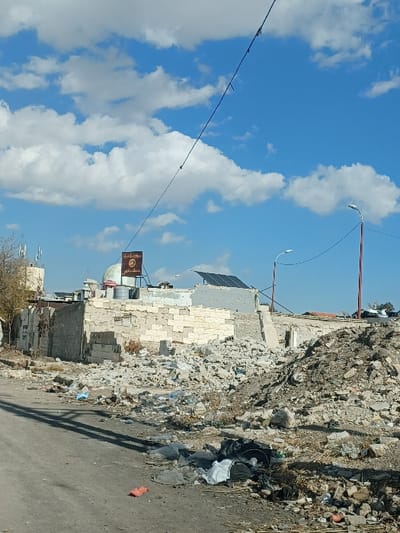 The image depicts a static urban scene in Al-Qadam Municipality, Syria, characterized by extensive demolition and accumulated rubble under a partly cloudy blue sky.

In the foreground, a worn asphalt road occupies the lower left, leading into the midground. Adjacent to the road, on the lower edge, assorted refuse including black and white plastic bags and an orange-red item are visible. To the right, a large, uneven mound of grey concrete and building rubble dominates, extending into the midground and containing fragments of masonry, rebar, and debris.

The midground features remnants of damaged structures. A partially intact building wall, constructed from light-colored stone or concrete blocks, exhibits signs of significant damage. Above this wall, a brown and white sign with Arabic script is mounted. Behind the sign, a light green dome is visible, possibly belonging to a religious structure or a water storage tank. Further right and slightly behind, a set of dark solar panels is installed on a flat, damaged rooftop. Overlapping the upper part of the scene, a thin black wire or cable with attached insulators stretches diagonally from the upper left towards the mid-right. Two modern-style streetlights with reddish-brown poles are positioned on the right side, one prominent and one partially obscured further back.

In the distant background, less distinct structures and the tops of telecommunication antennas on a building are visible on the far left. No individuals, animals, or active interactions are present in the frame. The overall environment suggests a state of disrepair, demolition, or post-conflict damage.