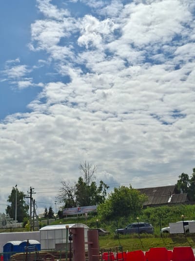 This is a vertically oriented, outdoor photograph taken during the daytime. The scene depicts a predominantly cloudy sky with patches of blue visible. The clouds are white and appear to be thin and wispy, suggesting a partly cloudy or overcast day.
In the background, there is a collection of buildings and trees. One building has a corrugated, weathered roof and appears to be older. Adjacent to it is a more modern structure with a white, reflective roof. A dark-colored car is parked on a grassy area. There are also several utility poles with wires crisscrossing overhead, indicating the presence of infrastructure.
To the right of the frame, there are rows of red stadium-style seats. Further into the scene, there are industrial-looking structures, possibly tanks or pipes, with a reddish-brown hue. A blue portable toilet is also visible in the foreground.
A banner with text is displayed on a white background, stretched between two poles. The text appears to be in Cyrillic script. One section of the banner shows what looks like a logo with red, white, and blue elements. The text is not entirely legible due to the image resolution and orientation. However, based on the provided location context, "Verhnij Uslon, Russia," it's plausible the banner contains information relevant to this area.
The overall impression is of a rural or semi-industrial setting, possibly near a public area or a construction site, under a cloudy sky. There are no people visible in the photograph. The scene does not convey any specific emotion and appears to be a straightforward documentation of the environment.