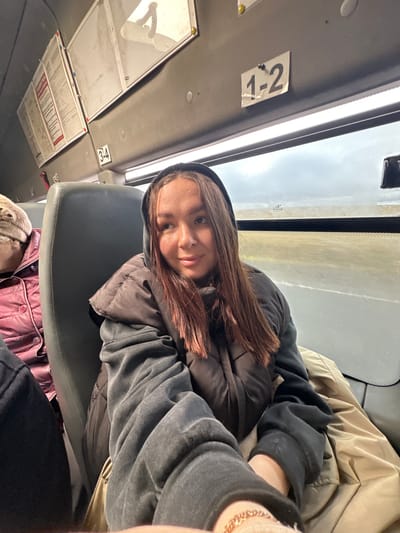 The image shows a woman sitting on a bus. She is wearing a dark gray hooded jacket and a gold chain bracelet is visible on her wrist. The bus has seat numbers 1-2 and 3-4 displayed on the ceiling. Through the window, a blurred landscape is visible. Another person wearing a red puffy jacket and a beige hat is partially visible in the foreground. There is no indication that this image was taken in Bol'shaya Sosnova, Russia.