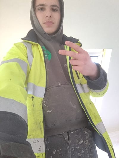 A single young male individual is depicted indoors in the city of Deerlijk, Belgium. The subject faces the camera directly, positioned from the chest up. He wears a dark grey hooded sweatshirt with the hood up, partially covered by an open bright yellow high-visibility jacket featuring horizontal grey reflective strips on the torso and arms. A green logo is visible on the left chest area of the grey sweatshirt. All visible clothing, including the subject's dark trousers, is soiled with white particulate matter. The subject's right hand is raised to chest height, forming a "V" gesture with the index and middle fingers extended. The background consists of a plain, unadorned white interior wall, with a bright light source visible in the upper right quadrant of the frame.