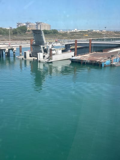 This image shows a dock and body of water in Loon-Plage, France. The photo captures the edge of a body of water, reflecting the structures and sky above. The structures appear to be industrial buildings and docks with metal beams and machinery. The water is a light green color, with ripples and reflections visible on the surface. The environment consists of an industrial area next to a body of water under a bright sky.