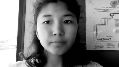 This is a black and white, close-up, shoulder-level portrait of a young East Asian woman. She is facing the camera directly, with her head slightly tilted. Her dark hair is loose and falls around her shoulders, with some strands framing her face. She is wearing a stud earring on her visible right ear.
Behind her and to her right, a portion of a document is visible. This document appears to be a schematic diagram of a bus route, with route numbers and names of locations in what seems to be a Cyrillic-based alphabet. Text on the document includes "№13 автобустук каттамдын кыймылынын СХЕМАСЫ" which translates to "Schematic of the movement of bus route No. 13". Other visible text includes location names like "Разаков к", "Ленин к", "Курманжан Датка", and numbered stops. There are also titles like "Аялдомела" (likely a place name or designation) and "Макулдашылды" (meaning "Agreed" or "Approved").
To her left, out of focus, is a bright window showing a glimpse of the outside. A white truck and a car are faintly visible through the window, suggesting the photo was taken during the daytime, possibly from inside a vehicle like a bus or car.
The woman's expression is neutral, and the overall mood of the image is calm. The lighting suggests it's a bright day, with the sunlight coming from the window. The setting appears to be urban or semi-urban, given the presence of the bus route map and vehicles outside the window.