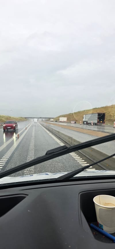 The image depicts a view from inside a vehicle, looking out through the windshield at a wet highway on a cloudy day. The road is marked with white lines and orange cones, indicating construction or maintenance. Several vehicles are visible on the highway, including a red car in the left lane and a large semi-truck with a trailer in the right lane. There are also some white portable structures, possibly temporary shelters or storage units, along the side of the road. In the foreground, the windshield wipers are partially visible. On the dashboard of the vehicle, a disposable coffee cup and a blue pen are present in a cup holder. The overall scene suggests a drive on a highway in or near Hatting, Denmark, under overcast and wet conditions.
