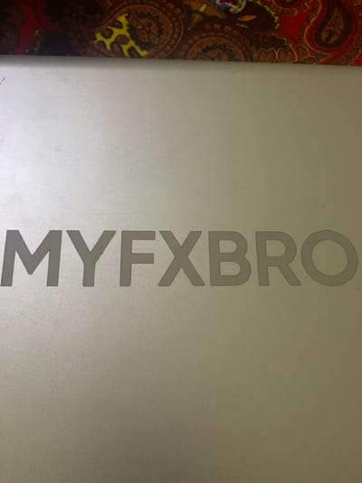 The image features the text "MYFXBRO" vertically aligned on a silver or light gray surface. The text is in a simple, sans-serif font and appears to be embossed or engraved into the surface. A small portion of a patterned fabric, possibly with paisley designs, is visible on the left side of the image, creating a contrast with the plain background. The lighting appears to be slightly diffused, giving the surface a smooth, matte appearance.