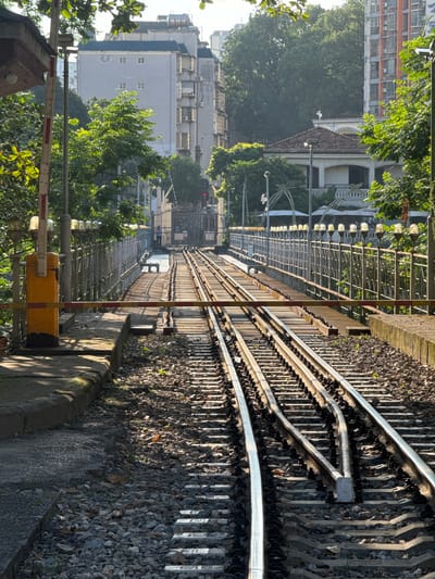 The image shows a railroad track in Lao Cai Ward, Vietnam. The tracks are surrounded by gravel. To the left, a pedestrian walkway is lined with a metal fence. Beyond the fence are buildings and trees. A yellow and red railroad crossing barrier is visible in the foreground on the left. The tracks converge in the middle distance.
