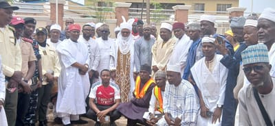 In Sabon Gari North, Nigeria, a group of people, primarily men, are gathered outdoors, posing for a photograph. The individuals are dressed in a mix of traditional Nigerian attire, including white and patterned kaftans, and some are wearing ceremonial caps and head wraps. Several men are in uniform, possibly representing security or official personnel. The scene appears to be a formal gathering, possibly an event or celebration, given the attire and the posed nature of the group. The background suggests an urban setting, with buildings and fencing visible.