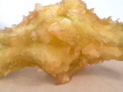 The image is a close-up, macro shot of a piece of fried food, likely a tempura vegetable, set against a plain white background. The food item is irregular in shape with a textured, crispy exterior that is golden brown in color. The light source appears to be from above and slightly to the left, casting soft shadows and highlighting the oily sheen of the fried batter. Small, lighter-colored pieces, possibly remnants of the vegetable, are visible within the batter. The focus is sharp on the food itself, with the background slightly blurred. There is no visible text in the image. The setting appears to be a food preparation or serving surface, given the stark white backdrop. The image is well-lit and the detail suggests it might be intended for a culinary context, perhaps a food blog, menu, or recipe illustration.