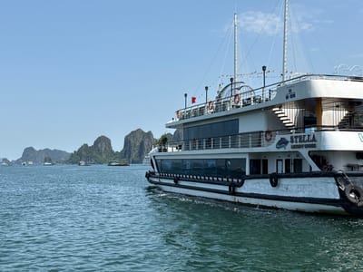 The image shows a large white tourist ship named "STELLA LUXURY CRUISE" on the water in Tuan Chau Ward, Vietnam. The ship is decorated with life preservers and flags. In the background, there are several small islands. The water is blue-green with small waves. The sky is blue with only a few clouds. Other smaller boats are visible in the distance.