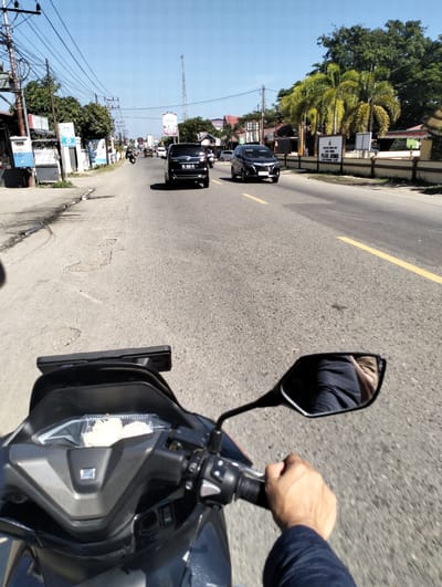 A first-person perspective shows a person riding a motorcycle on a road in Meunasahkota, Indonesia. The rider's hands are on the handlebars, and their navy blue sleeve is visible. In the motorcycle's mirror, a reflection of the rider's face and navy blue clothing is visible. In a compartment on the motorcycle, a clear plastic container holds two white food items. The road is asphalt with a yellow dividing line. On the left side of the road, there are utility poles, electrical wires, buildings, and trees. On the right side of the road, there are palm trees, a building with a sign that reads "POLISI JATENG" (Police Central Java), and other structures. Two black cars are traveling in the same direction as the motorcycle, and other vehicles are visible in the distance. The sky is clear and blue.