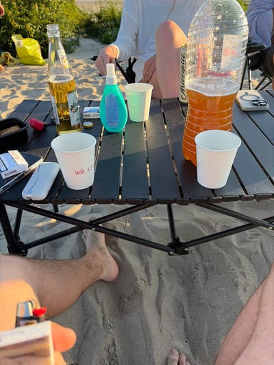 This is a medium shot, eye-level, outdoor photograph taken in the city of Povarovka, Russia. The scene is on a sandy beach during daytime, with lush green foliage visible in the background. A foldable black table is set up on the sand, displaying various items.
On the table, there is a tall, clear plastic bottle partially filled with an amber liquid, possibly beer or a soft drink. Next to it is a glass bottle of beer, with condensation on its surface and some foam at the top. There are two white paper cups, one with red Cyrillic text that appears to read "мой спорт" (my sport). A turquoise colored bottle of what looks like hand sanitizer or soap is also present. To the left, a pack of cigarettes labeled "HEETS" and a phone are on the table, along with a small black container filled with a granular substance. A red object, possibly a lighter or a tool, is also visible.
In the foreground, a person's bare legs and feet are partially visible, covered in sand. The sand appears coarse and light in color. The overall atmosphere suggests a relaxed outdoor gathering.