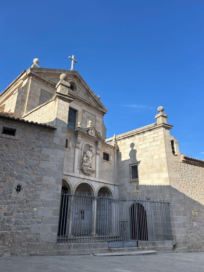 The image shows a stone building against a bright blue sky. The building has a cross on top, a statue in a niche, and a metal grate over an arched entryway. The building is located in the city of Ávila, Spain.