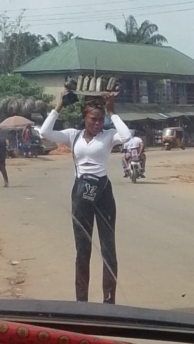 This image captures an outdoor street scene in Igbo Ukwu, Nigeria, centered on a young adult female street vendor. The woman, appearing light-skinned with dark hair, stands in the foreground, directly facing the camera. She wears a white long-sleeved crop top with a V-neck and a midriff cutout, dark trousers, and a black cross-body bag featuring a prominent white "LV" style logo. Her hands are raised, supporting a flat, rectangular wooden tray balanced on her head, which holds several oblong, light-colored packaged items.

The background shows an unpaved or dirt road surface. In the mid-ground, a person rides a white motorcycle towards the right. To the far right, a yellow motorized tricycle is visible. The left side of the mid-ground contains indistinct figures and vehicles near a red or orange umbrella. The central background is dominated by a multi-story building with a green corrugated metal roof, partially obscured by green foliage, including palm trees. Overhead utility lines are visible against the light sky. The photograph is taken from a low angle, with a portion of a vehicle's interior, including a red patterned element, visible at the very bottom of the frame.