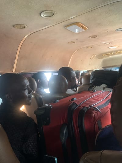 The image shows the interior of a passenger van, likely a commercial bus, with several individuals seated inside and luggage packed around them. The ceiling of the van features overhead lighting and air vents. The passengers appear to be of African descent. One passenger in the foreground is seen in profile, looking forward. A large red suitcase is prominently placed in the aisle, alongside other dark-colored luggage. The scene suggests travel, possibly a long-distance journey. The image context provided states the location is Elelenwo, Nigeria.