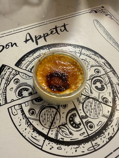 A crème brûlée in a clear glass ramekin sits on a placemat with a black and white drawing. The placemat has text that says "Bon Appétit" and a drawing of a knife. The drawing underneath the ramekin appears to be a stylized depiction of a pizza with gears and mechanical elements. The crème brûlée has a perfectly caramelized, golden-brown sugar crust, with some areas appearing slightly darker, indicating a rich caramelization. The setting is likely a home kitchen or a casual dining establishment in Oakville, Canada, given the provided context, although no specific landmarks or identifying features of Oakville are visible in the image itself. The lighting suggests indoor ambient light.