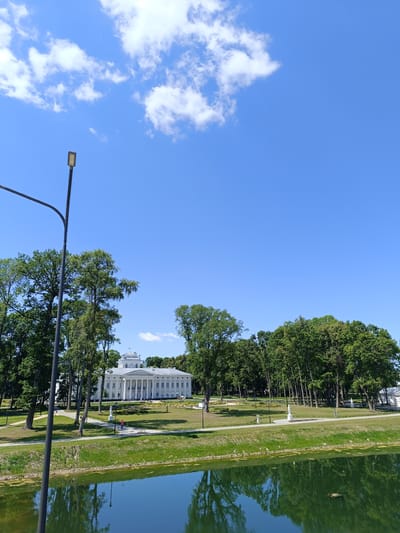 A wide shot captures the facade of a large, white classical building with columns and a dome, set against a backdrop of lush green trees under a bright blue sky with scattered white clouds. In the foreground, a body of water reflects the trees and sky. A black lamppost stands on the left side of the frame, partially obscuring the view of the building and trees. The grounds in front of the building are manicured with lawns and pathways, and a few small figures, possibly people, can be seen walking on the paths. The scene is peaceful and suggests a well-maintained park or estate. The location is identified as Добасненскі сельскі Савет, Belarus.