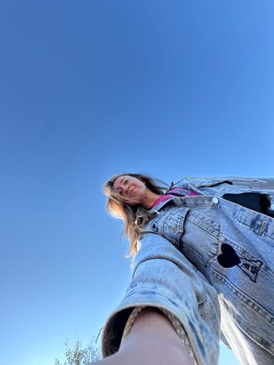 A low-angle selfie, taken in Ust'-Silayka, Russia, features a single individual, likely female, positioned in the upper right portion of the frame. The subject has light brown hair, which is partially illuminated by sunlight, and a light-toned complexion, displaying a slight smile while looking directly downwards towards the camera. She is wearing a faded blue denim jacket with visible stitching and two distinct patches: a black heart shape and, beneath it, a white letter 'A' with a black outline, both located on the right side of the jacket. A pink or magenta t-shirt is visible underneath the open jacket. The individual's right arm, likely holding the camera, extends from the bottom right into the frame. The background is dominated by a clear, bright blue sky. Minimal green foliage is visible at the extreme bottom left edge of the image.