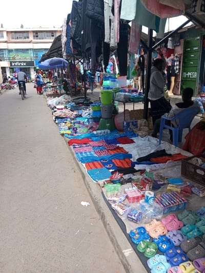 The image depicts a bustling outdoor market situated along a paved street in Ildamat ward, Kenya.

On the right side of the image, numerous vendor stalls are arranged along the street, extending into the background. These stalls are covered by dark tarpaulins and supported by metal frames. Merchandise is displayed on white ground tarps, on tables, and hanging from overhead racks.

In the immediate foreground on the right, a variety of goods are presented on a white tarp. These include neatly arranged rows of multi-colored children's plastic clogs (e.g., pink, blue, green, grey, orange), packaged toothbrushes, rolls of toilet paper, and assorted small boxed and plastic-wrapped items. Further back at the stalls, folded patterned textiles (red, blue, checkered) are visible, along with stacks of plastic buckets (green, blue, grey) and clear plastic containers. Overhead, various articles of clothing, including jackets and towels, hang from racks. A blue market umbrella is also open over one of the stalls in the mid-ground.

Two individuals are present near a stall on the mid-right: one person, wearing a light grey hoodie and a dark skirt, stands facing left. Another individual, viewed from behind, sits on a blue plastic chair, wearing a grey hoodie. In the far background on the right, a green shipping container-like structure with "M-PESA" branding is visible, positioned adjacent to mannequins displaying clothing.

The left side of the image features a grey paved street with a visible curb separating it from the market. In the mid-ground, a person on a bicycle, wearing a dark shirt and light trousers, cycles away from the camera. In the background on the left, multi-story buildings are visible, including one with a prominent green and white "Infinix" sign. Several other pedestrians are observed in the distance among the market stalls and along the street. The overall scene indicates active commerce in an urban environment.